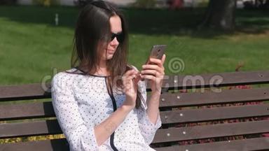 年轻的黑发女人在<strong>公园里坐在</strong>长凳上的手机上键入一条信息。